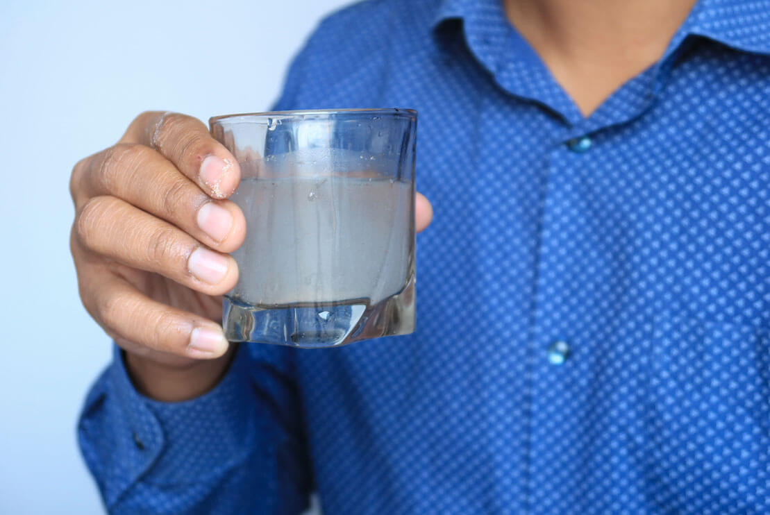 A man in a blue dotted shirt holding a glass of dirty water